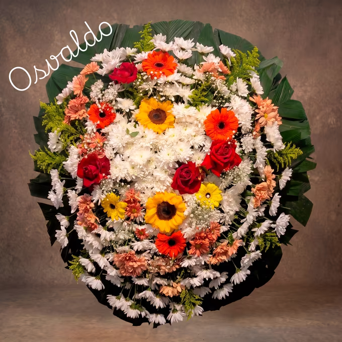 Coroa oval de crisântemos brancos com girassóis, gérberas laranja e salmão e rosas vermelhas sobre folhas verdes de palmeira.