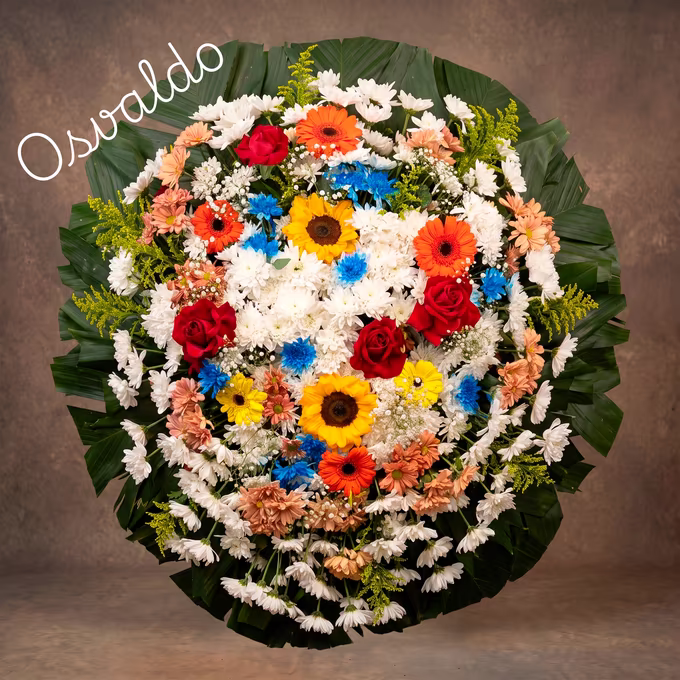 Coroa oval de crisântemos brancos e salmão, girassóis, gérberas laranja, rosas vermelhas e flores azuis sobre leque de folhas de palmeira.