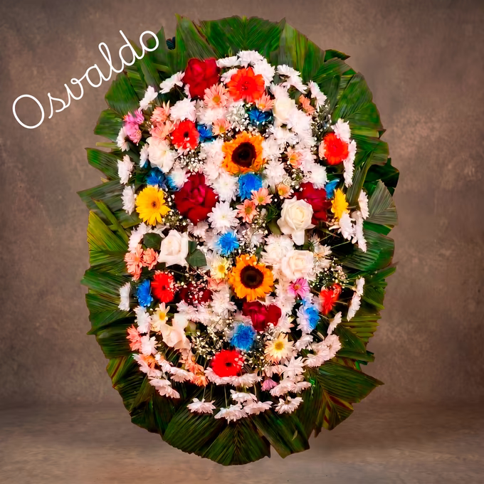 Coroa oval com crisântemos brancos e salmão, gérberas vermelhas, laranja e amarelas, girassóis, rosas creme e crisântemos azuis sobre folhas de palmeira.